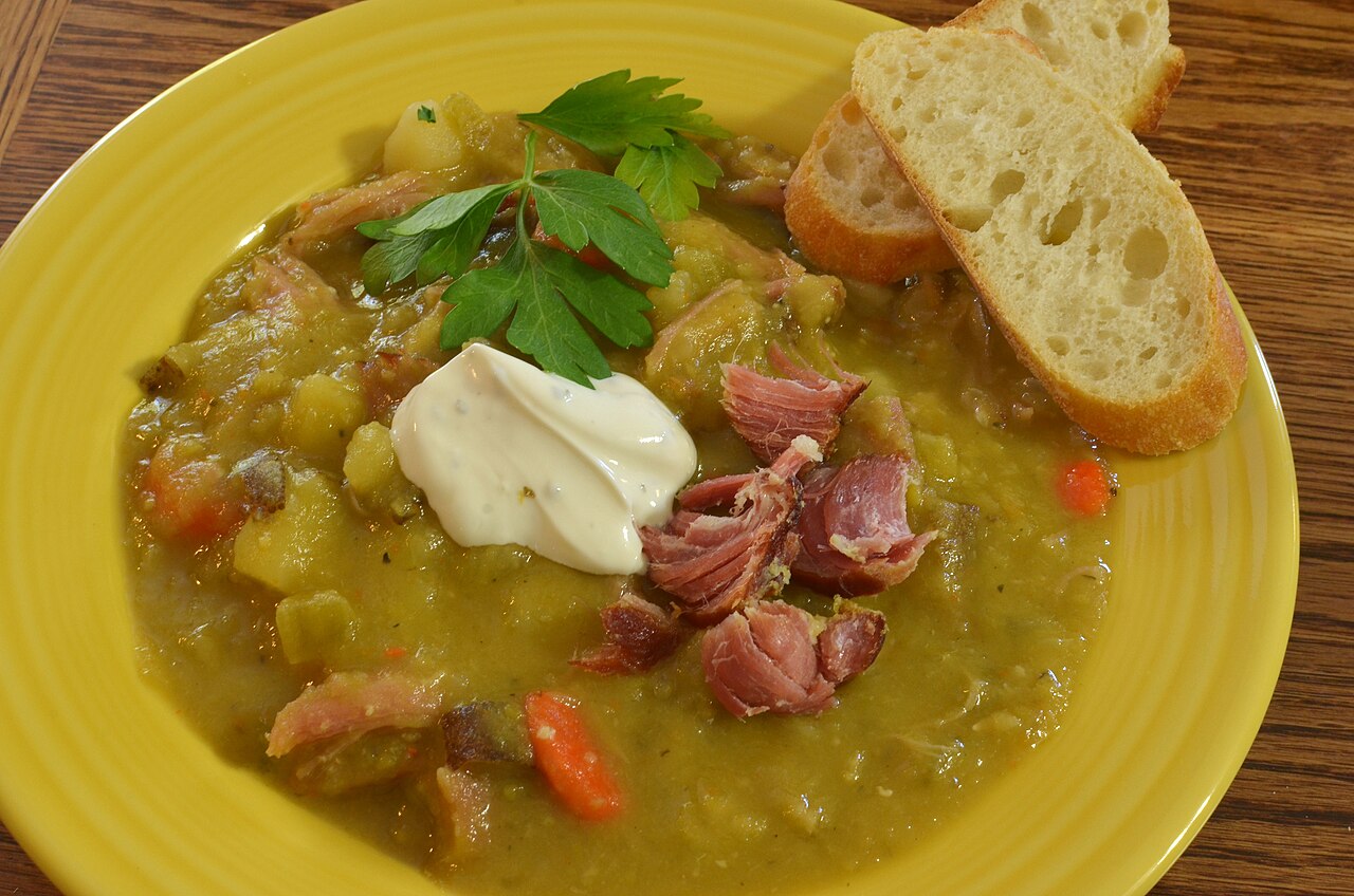 Thick split pea soup with shredded ham, diced vegetables, a spoonful of cream and parsley in a yellow bowl with slices of bread photo_0 Thick split pea soup with shredded ham, diced vegetables, a spoonful of cream and parsley in a yellow bowl with slices of bread photo_0